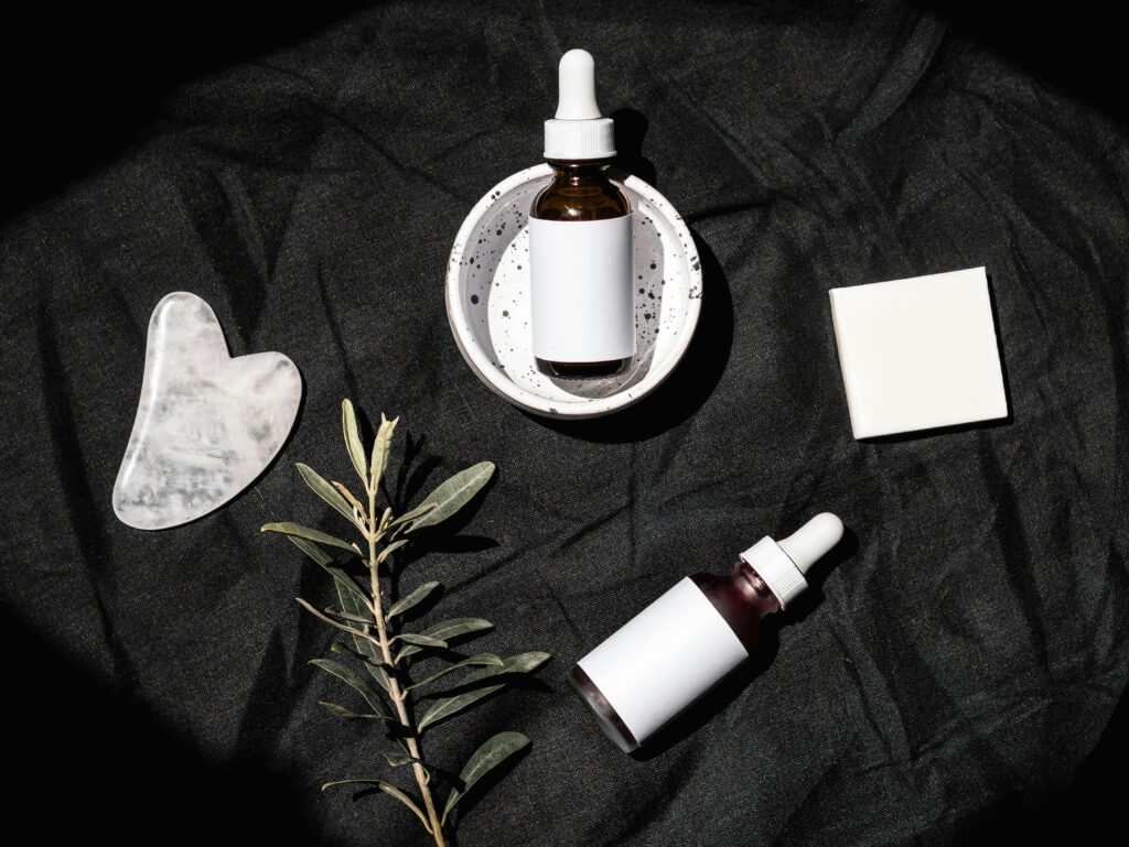 Stylish flat lay of skincare serum bottles with gua sha tool and leaves on a dark textile background.